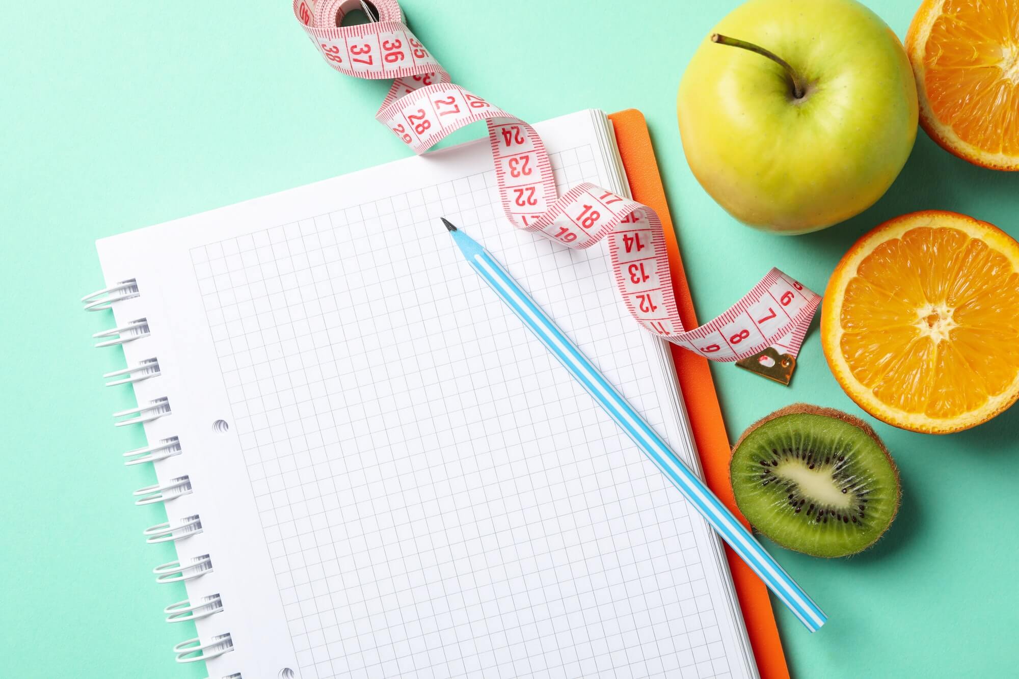 Blank notebook, pencil, fruit, and measuring tape on a mint-colored surface