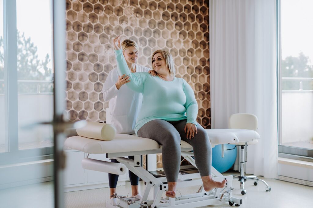 Mid adult physiotherapist woman exercising with overweight woman indoors in rehabilitation center