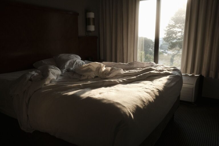 Unmade bed with soft white duvet by a large window with warm morning sunlight, representing the sleep, hormone, and mood connection