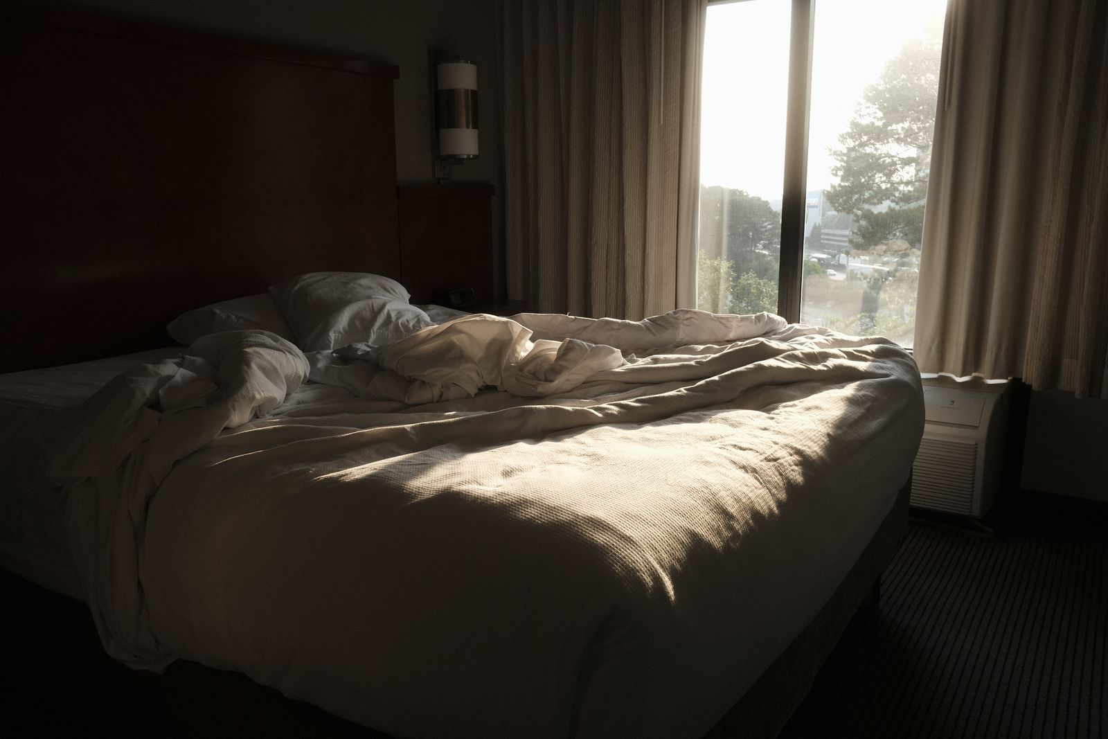 Unmade bed with soft white duvet by a large window with warm morning sunlight, representing the sleep, hormone, and mood connection