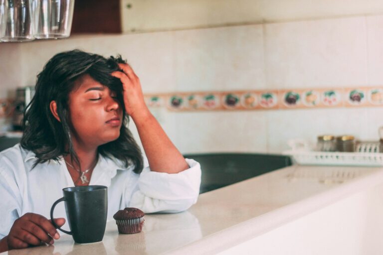 Black woman in white robe with morning coffee, hand on forehead in fatigue, in bright Florida kitchen