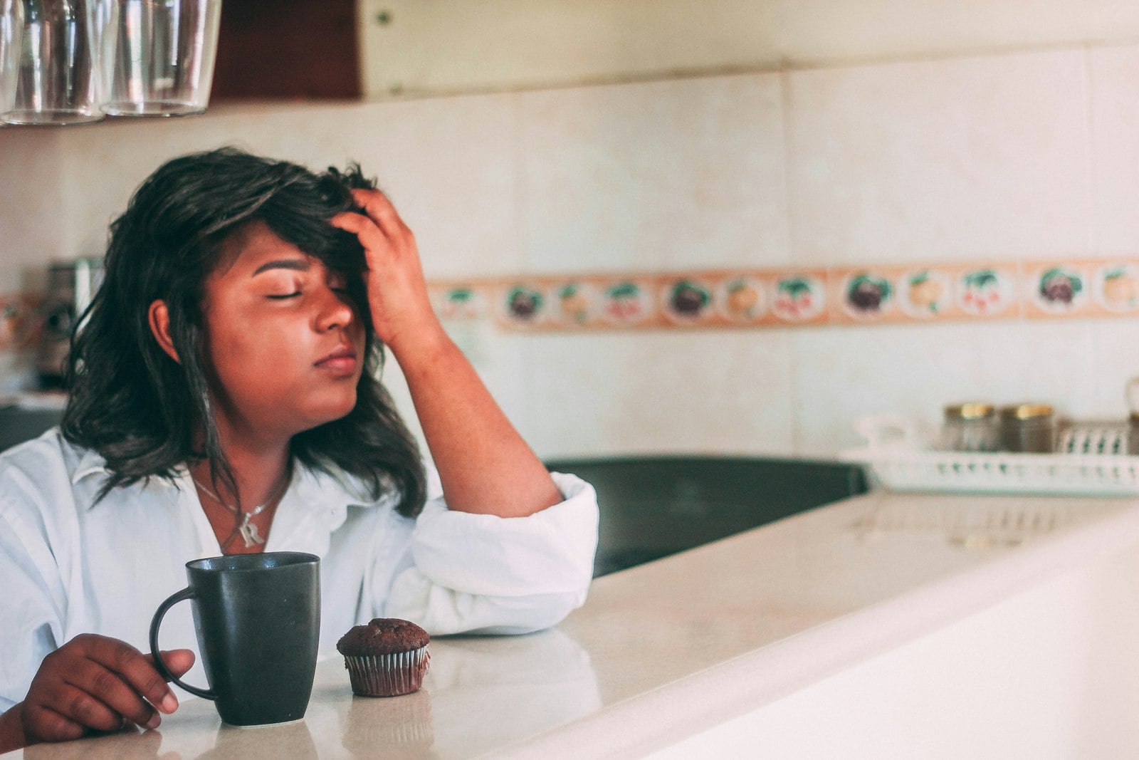 Black woman in white robe with morning coffee, hand on forehead in fatigue, in bright Florida kitchen