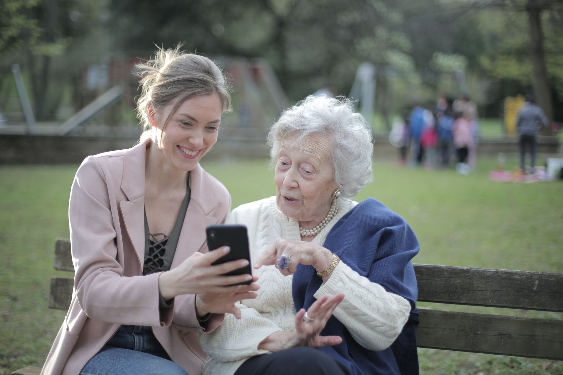 An older woman and a younger woman looking at a smartphone together on a park bench.